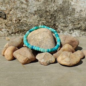 Peut inclure: Un bracelet en pierres précieuses turquoise et bleu composé de petites perles rondes. Le bracelet est posé sur un tas de pierres brunes.