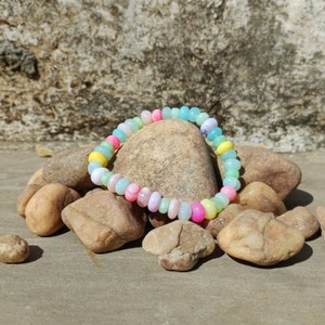 Peut inclure: Un bracelet coloré en perles avec des nuances pastel de bleu, vert, rose et jaune. Le bracelet est composé de petites perles rondes et est posé sur un tas de pierres.