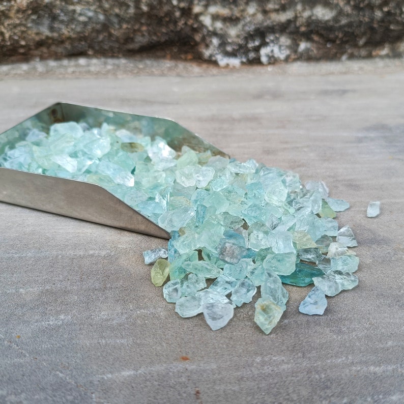 Puede incluir: Una pala de metal llena de virutas de piedra preciosa de aguamarina. Las virutas son de color azul claro y est&aacute;n dispersas sobre una superficie gris.