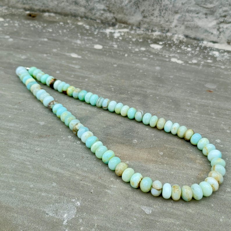 May include: A necklace composed of round, light blue and tan beads. The beads are strung together to form a necklace. The necklace is displayed on a gray surface. The beads have a polished appearance.