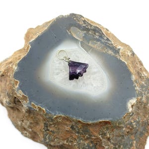 May include: A faceted, dark purple gemstone pendant with a silver bail. The pendant is displayed on a gray and white agate slice with a brown, textured exterior. The gemstone has a unique, irregular shape.