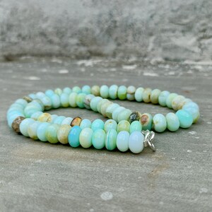 May include: A necklace composed of oval-shaped, light blue and cream-colored beads. The beads are strung together, forming a circular shape. The necklace has a silver-colored clasp. The beads have a polished, smooth appearance.