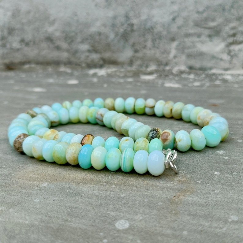 May include: A necklace composed of oval-shaped, light blue and cream-colored beads. The beads are strung together, forming a circular shape. The necklace has a silver-colored clasp. The beads have a polished, smooth appearance.