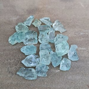 Puede incluir: Una colecci&oacute;n de virutas de cristal de aguamarina azul claro. Las virutas son de varios tama&ntilde;os y formas, algunas tienen una forma m&aacute;s puntiaguda y otras son m&aacute;s redondeadas. Las virutas est&aacute;n dispersas sobre una superficie gris.