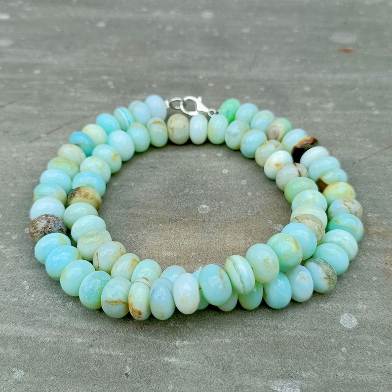 May include: A necklace composed of light blue and green oval-shaped beads. The beads are strung together, forming a double strand. The necklace is secured with a silver-colored clasp. The beads have a natural, organic appearance.