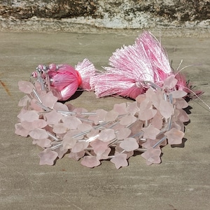 May include: A strand of pink rose quartz beads in the shape of stars, strung on silver wire. The beads are arranged in a loose pile, with a pink tassel hanging off the side.