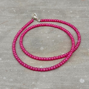 May include: A bright pink beaded necklace with a silver clasp. The necklace is made of small, round beads and is displayed on a gray surface. The necklace is a delicate piece of jewelry.