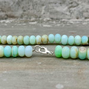 May include: A close-up of a necklace featuring light blue and green beads. The beads are oval-shaped and strung together with a silver clasp. The necklace is displayed against a neutral background.