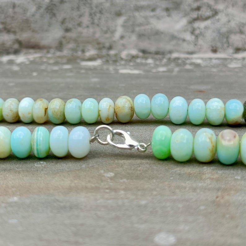 May include: A close-up of a necklace featuring light blue and green beads. The beads are oval-shaped and strung together with a silver clasp. The necklace is displayed against a neutral background.