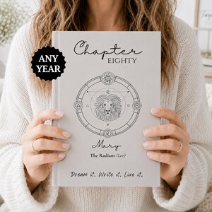 Puede incluir: Un libro de tapa dura gris claro con el texto "Chapter EIGHTY" y "ANY YEAR" en una insignia circular negra. La portada muestra una ilustración central de un león dentro de un círculo zodiacal geométrico, el nombre "Mary", "The Radiant (Leo)" y "Dream it. Write it. Live it."