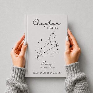 Puede incluir: Un libro de tapa dura blanco con texto negro y un gráfico de constelación. La portada dice Chapter EIGHTY, Mary, The Radiant (Leo) y Dream it. Write it. Live it. El libro es sostenido por dos manos con suéteres de punto gris.