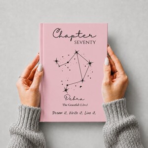 May include: A light pink hardcover journal featuring the black text Chapter SEVENTY, a black Libra constellation graphic, the name Debra, the text The Graceful (Libra), and the phrase Dream it. Write it. Live it. printed on the front cover.
