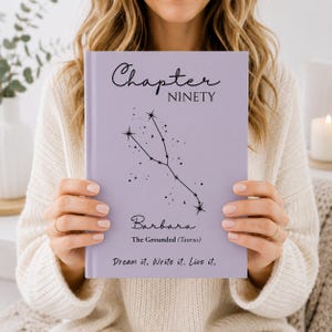 Könnte beinhalten: Ein hellviolettes Notizbuch mit festem Einband, das den Text Chapter NINETY, den Namen Barbara, den Zusatz The Grounded (Taurus) und das Zitat Dream it. Write it. Live it. zeigt. Ein schwarzes Sternbild-Liniengrafik ist mittig platziert.