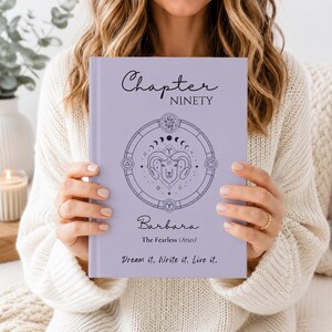 Può includere: Un diario con copertina rigida color viola chiaro con il testo Chapter NINETY, Barbara, The Fearless (Aries) e Dream it. Write it. Live it. La copertina presenta un'illustrazione circolare dello zodiaco viola scuro con un ariete al centro.