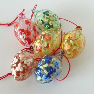 Könnte beinhalten: Sechs gläserne Eier-Anhänger mit Blumenmustern. Die Anhänger sind in verschiedenen Farben bemalt, darunter Rot, Orange, Gelb, Grün, Blau und Weiß.