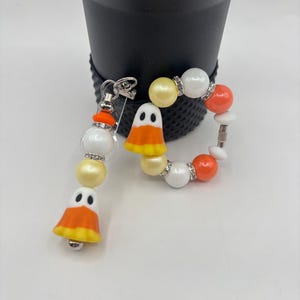 Op de afbeelding: Een Halloween-thema sleutelhanger en armband set. De sleutelhanger heeft een candy corn spook bedel met witte, oranje en gele kralen. De armband heeft witte, oranje en gele kralen met een candy corn spook bedel. Beide hebben zilveren accenten.