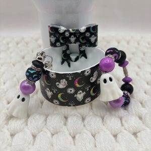 May include: A black silicone tumbler boot featuring a pattern of white ghosts, colorful crescent moons, and mushrooms. The boot is accessorized with a black bow and two beaded charms, each featuring a white ghost bead, purple beads, and black beads.