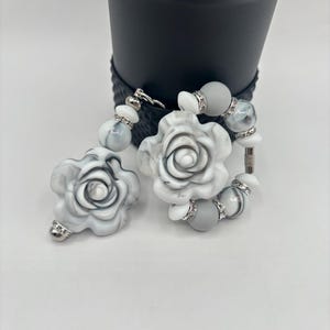 May include: A pair of white and grey rose-shaped earrings with silver accents. The earrings feature a marbled design and are adorned with small, sparkling rhinestones. The earrings are connected by a silver chain and are displayed against a neutral background.