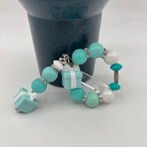 May include: A beaded bracelet with turquoise, white, and silver accents. The bracelet features round beads and small gift box charms. The beads are strung on a clear cord. The bracelet is next to a dark teal tumbler.