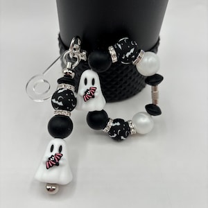 May include: A Halloween-themed keychain and bracelet set. The keychain features black and white patterned beads, a ghost charm with a bat design, and a silver clasp. The bracelet has similar beads and ghost charms.