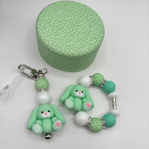 Könnte beinhalten: Eine hellgrüne runde Schachtel mit floralem Muster, ein Schlüsselanhänger und ein Armband. Der Schlüsselanhänger und das Armband zeigen hellgrüne Hasenanhänger mit rosa Herzen und weißen und grünen Perlen. Die Schachtel hat einen Durchmesser von etwa 10 cm.