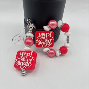 以下が含まれることがあります： 「Yep! Still Single」の文字が入った赤いキーホルダー、ブレスレット、チャーム。キーホルダーは透明なプラスチックのループと赤いビーズが付いています。ブレスレットは赤、ピンク、白のビーズが特徴です。チャームは白い文字が入った赤い正方形です。