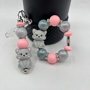 May include: A beaded keychain and bracelet set with grey cat charms, pink and grey beads, and silver accents. The keychain has a clear plastic attachment. The bracelet has a silver clasp.