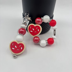 May include: A red and white beaded keychain and bracelet set. The keychain features a heart-shaped cookie charm with white sprinkles. The bracelet and keychain have red and white beads with rhinestone accents.