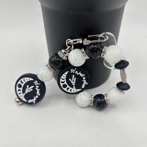 May include: A black and white beaded accessory with a silver clasp. The accessory features black and white speckled beads, clear rhinestone spacers, and two round black charms with a crescent moon and text that reads "It's just a phase."
