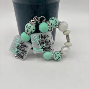 Peut inclure: Un ensemble de porte-clés et de bracelets avec des accents vert menthe et argentés. Le porte-clés comporte deux breloques carrées avec un motif d'ancre et le texte "Hope is an Anchor for the Soul". Le bracelet comprend des perles nacrées et vert menthe.