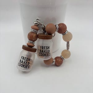 May include: A keychain featuring two miniature glass jars with brown lids, each labeled "FRESH BAKED COOKIES" in black text. The jars are connected by a beaded strand with brown, tan, and silver beads.