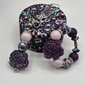 May include: A purple beaded bracelet with floral accents and a matching floral-patterned box. The bracelet features large purple flower-shaped beads, light pink beads, and decorative beads with silver accents. The box has a black background with a floral pattern.