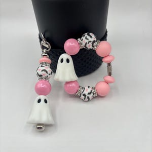 May include: A pink and white beaded keychain and bracelet set. The keychain features a white ghost charm, pink beads, and beads with a black and white pattern. The bracelet has similar beads and a ghost charm. The set is designed with silver accents.