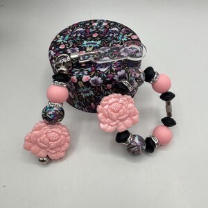 May include: A set of baby accessories, including a pacifier clip and a bracelet, featuring pink and black beads, floral accents, and a floral-patterned container. The pacifier clip has a pink flower and a silver clip.