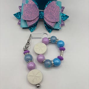 May include: A mermaid-themed set featuring a glittery purple and teal hair bow, a beaded bracelet, and a keychain. The bracelet and keychain include light blue, purple, and white beads, with sand dollar accents. The bow is layered with glitter and patterned teal fabric.