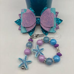 May include: A mermaid-themed accessory set featuring a large bow with glittery purple and teal accents, and a beaded keychain and bracelet with starfish charms. The set includes shades of purple, teal, and blue.