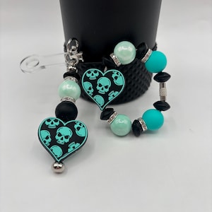 Może przedstawiać: Zestaw breloczka i bransoletki z zawieszką w kształcie serca z turkusowym wzorem czaszki na czarnym tle. Breloczek ma turkusowe i czarne koraliki oraz zapięcie na agrafkę. Bransoletka ma turkusowe i czarne koraliki ze srebrnymi akcentami.