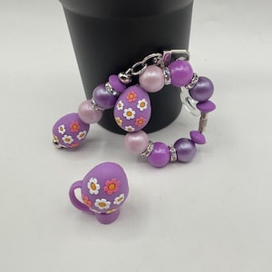 May include: A purple beaded bracelet and matching miniature teacup. The bracelet features various sized beads in shades of purple and pink, with floral egg-shaped beads. The teacup is purple with white and orange flower details.