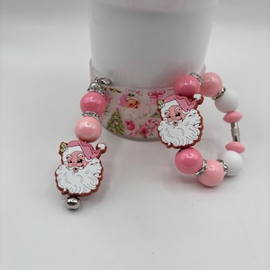 May include: Pink and white beaded bracelet and charm featuring a Santa Claus design. The bracelet has pink, white, and silver beads. The Santa Claus charm has a pink hat and a white beard.