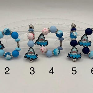 Puede incluir: Una colección de seis pulseras y llaveros con temática de tiburones. Cada uno presenta un dije de tiburón, con cuentas azules y blancas. Los llaveros y pulseras son de varios tonos de azul, con detalles metálicos plateados.