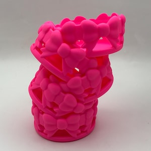 May include: A stack of three bright neon pink silicone bands, each featuring a repeating pattern of raised bows connected in a continuous loop. The material is flexible and matte, with a uniform vibrant color throughout the entire structure.