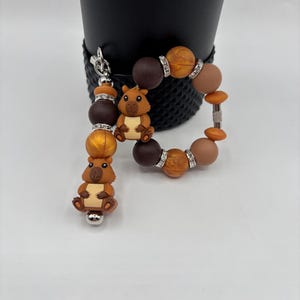 May include: A beaded keychain and bracelet set featuring brown, orange, and gold beads. The keychain and bracelet both have a small, brown, cartoon-style beaver charm. The beads are accented with silver-colored spacers.