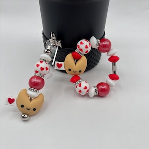 May include: A keychain and bracelet set featuring a fortune cookie charm and red and white heart-patterned beads. The keychain has a silver clasp and a small tag with a heart. The bracelet has a similar design with a fortune cookie charm.