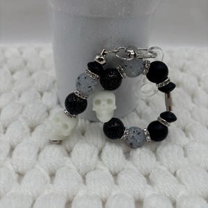 May include: A circular keychain featuring two white skull beads, black glittery beads, matte black beads, and grey speckled beads. The beads are separated by small silver-toned spacers encrusted with clear rhinestones, and the piece includes a silver metal clasp.