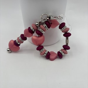 以下が含まれることがあります： ピンクとバーガンディのビーズブレスレットとキーホルダーのセット。ブレスレットはハート型のビーズとラインストーンアクセントの丸いビーズが特徴です。キーホルダーは、ハート型のビーズとシルバーカラーの留め金が付いた同様のデザインです。