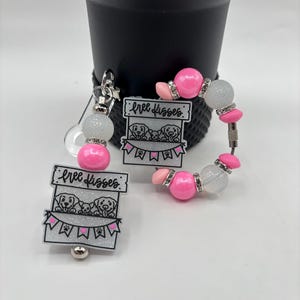 May include: A set of two accessories: a retractable badge reel and a beaded bracelet. Both feature a design with the text "Free Kisses" above an illustration of three puppies. The accessories have pink, white, and silver accents.
