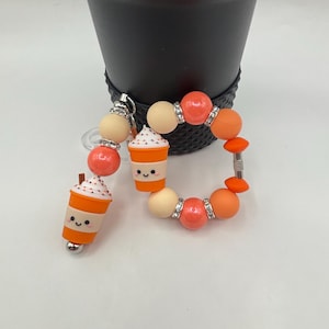 Może przedstawiać: Zestaw breloczka i bransoletki z motywem jesiennym. Breloczek ma mały, pomarańczowo-biały charms w kształcie filiżanki kawy. Bransoletka ma pomarańczowe, kremowe i dżetowe koraliki. Przedmioty znajdują się na białej powierzchni.