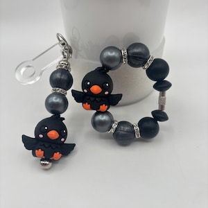 Op de afbeelding: Een zwarte en oranje vogel-thema sleutelhanger en armband set. De sleutelhanger heeft een veiligheidsspeld sluiting en een vogel bedel met grijze kralen. De armband heeft zwarte en grijze kralen met strass accenten en een vogel bedel.