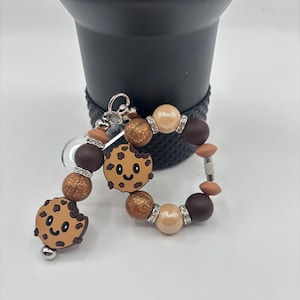 May include: A beaded keychain with a cookie theme. The keychain features brown, tan, and glittery beads, along with two smiling cookie charms. The keychain is attached to a silver clasp.