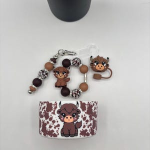 Könnte beinhalten: Eine Sammlung von Accessoires mit einem Highland-Kuh-Design. Enthält eine Hülle mit braunem und weißem Muster, einen Schlüsselanhänger mit Perlen, ein Armband mit Perlen und einen kleinen Anhänger. Die Kuh ist braun mit schwarzen Hörnern und einem niedlichen Ausdruck.
